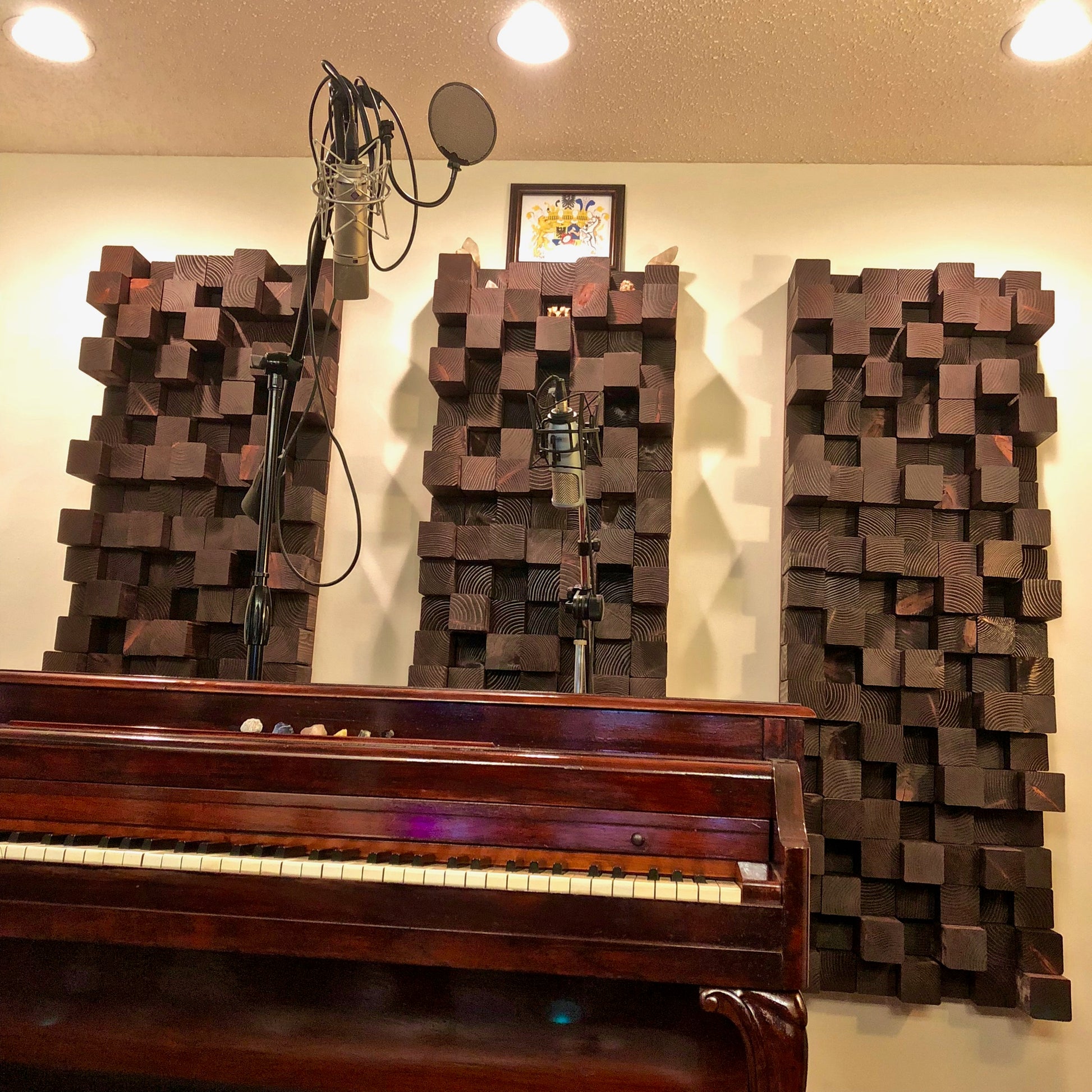 Three dark brown wooden 3D block wall art panels mounted on a beige wall above a dark wood upright piano, with two studio microphones and stands in front of the panels.
